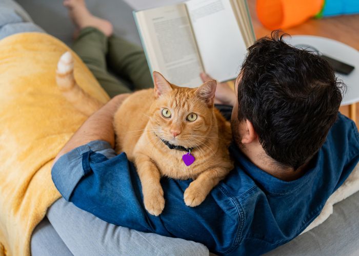 Man sitting on a couch reading a book with his cat on his shoulder.